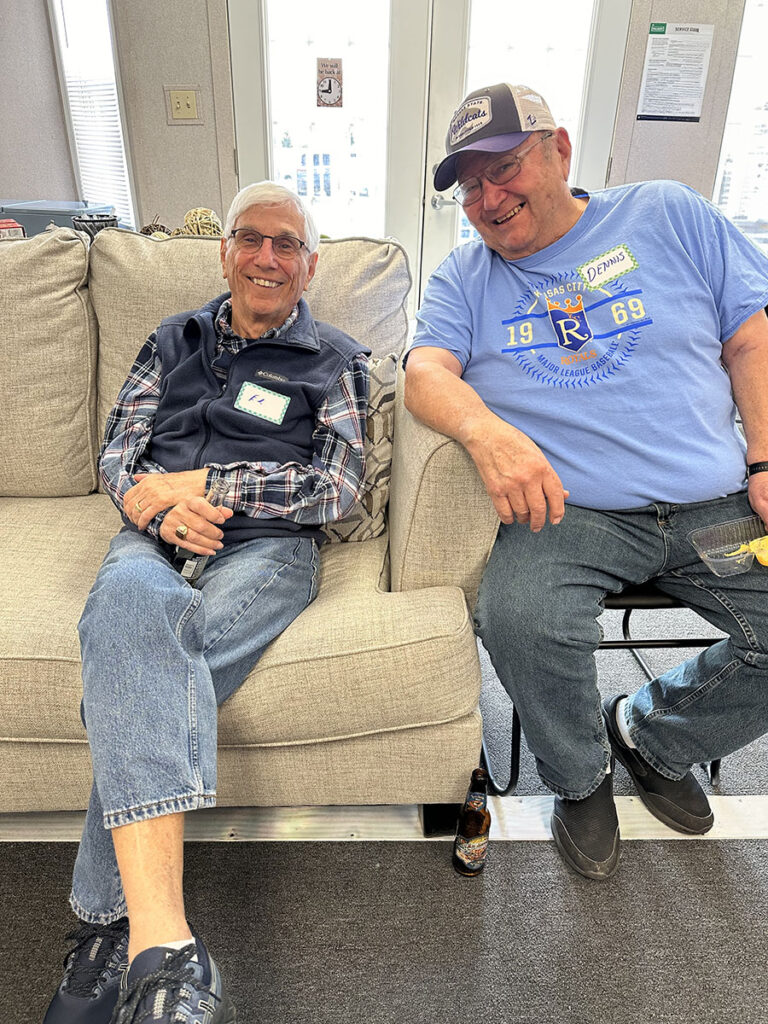 Two vibrant older men relaxing on a couch, sharing laughter, beers, and a tasty snack.