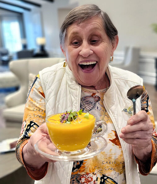 Smiling senior resident joyfully holds a bowl of vibrant orange soup garnished with greens, wearing a colorful butterfly-patterned shirt and white vest, ready to enjoy the meal.