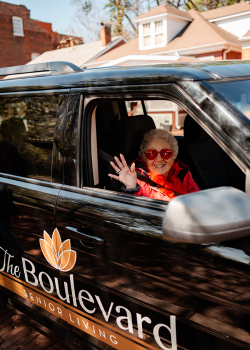 Senior resident wearing heart-shaped sunglasses waves happily from the passenger seat of a 'The Boulevard Senior Living' vehicle, showcasing an enjoyable outing on a sunny day.