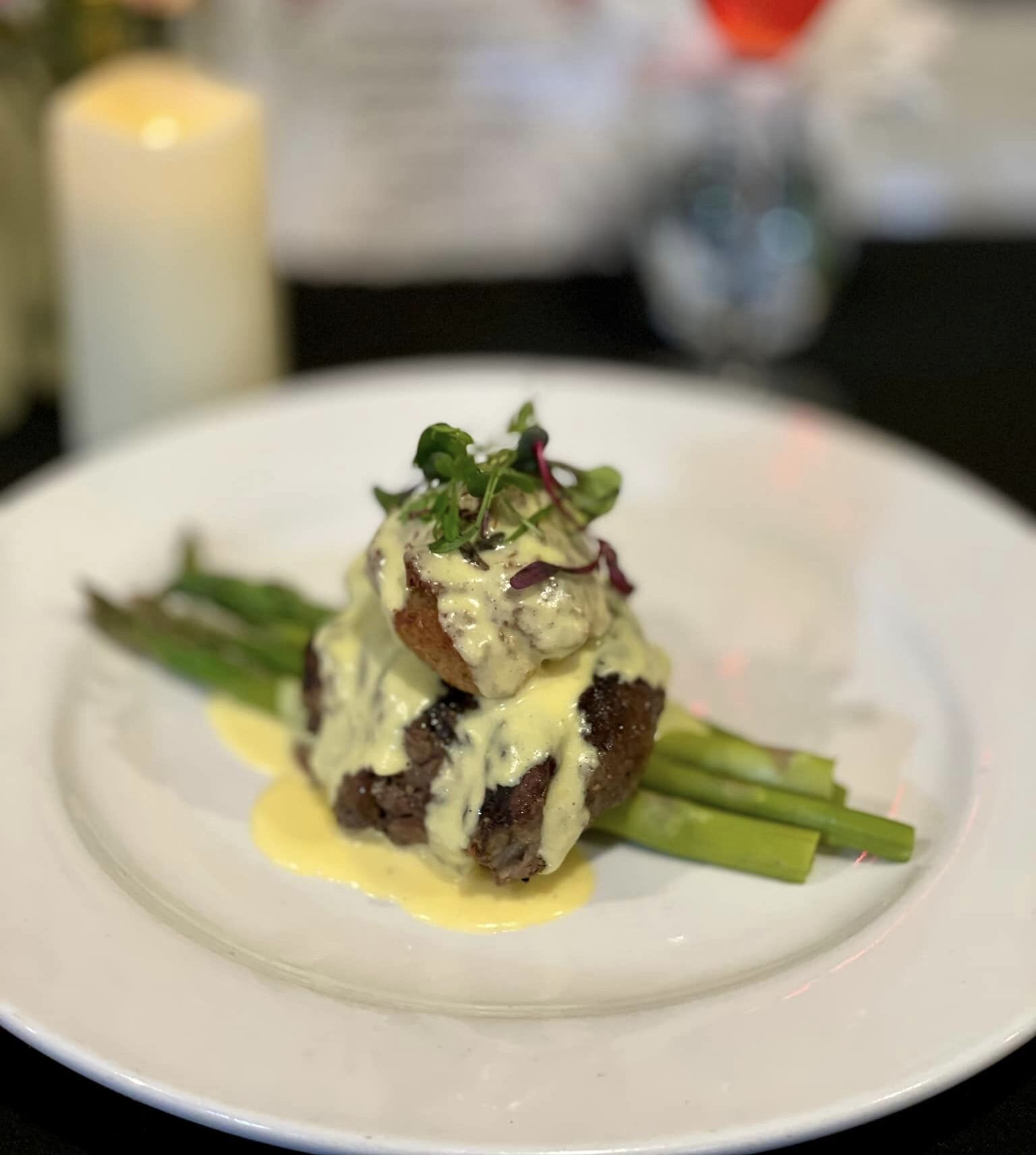 Plated gourmet steak with asparagus, topped with creamy hollandaise sauce and garnished with microgreens, set against a blurred background of a candle, flowers, and drink.