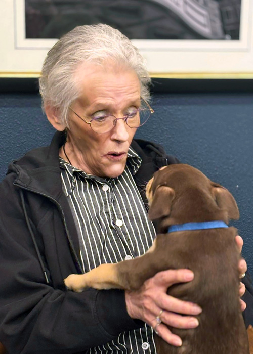 A senior resident smiles while holding a brown puppy during a joyful pet therapy visit.