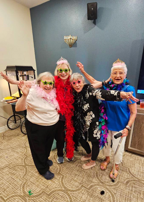 Four senior residents strike playful poses in boas, tiaras, and star glasses during a themed dress-up event celebrating Gorgeous Grandma Day at the community.