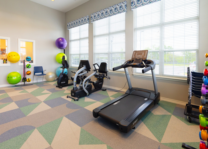 A bright and inviting wellness room with large windows, fitness bikes, a treadmill, exercise balls, and hand weights arranged neatly on racks.
