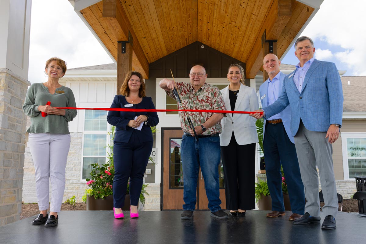 A group of six individuals stands under a wooden portico during a ribbon-cutting ceremony at The Crestone Senior Living, with one man cutting the red ribbon.