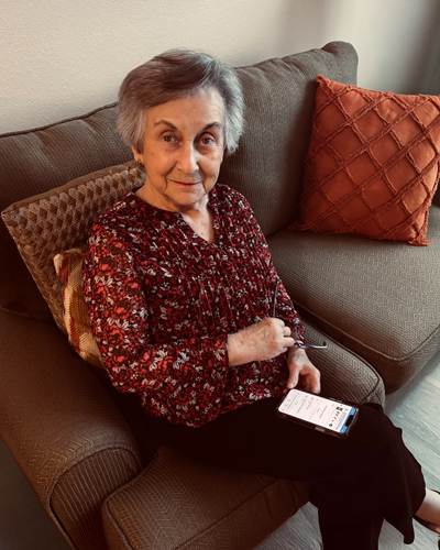 A senior woman with short gray hair sits on a couch holding a smartphone. She wears a red floral blouse and smiles warmly at the camera.
