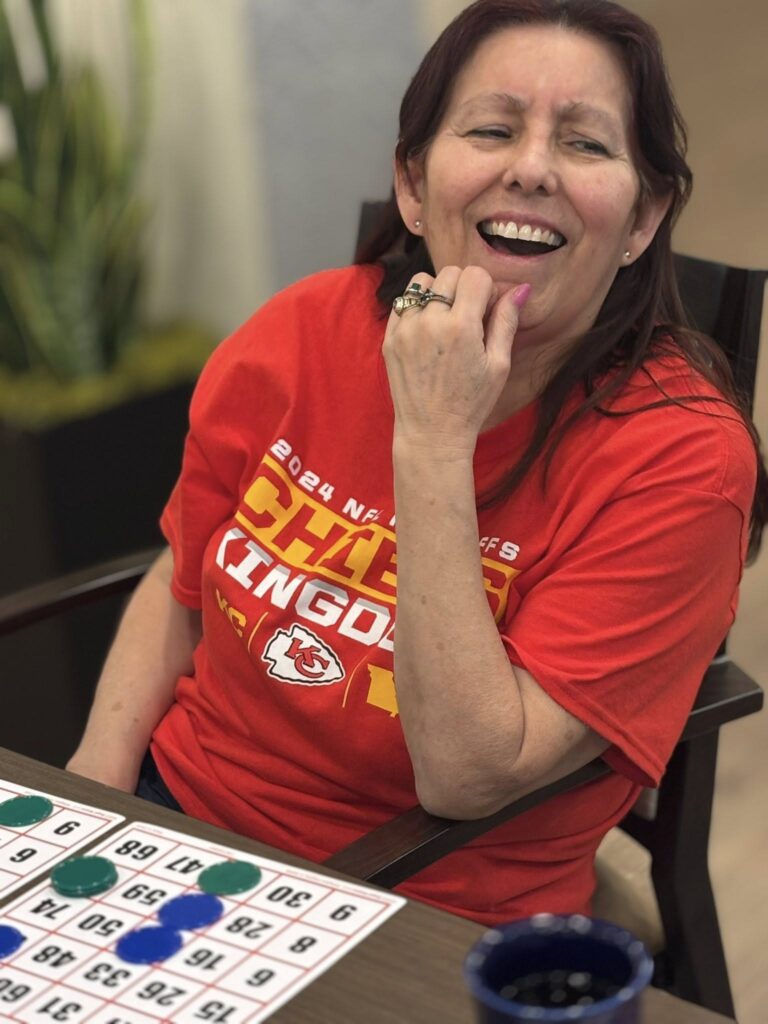 A smiling resident in a bright red Kansas City Chiefs shirt laughs while playing a lively game of bingo, enjoying community fun and friendly competition.