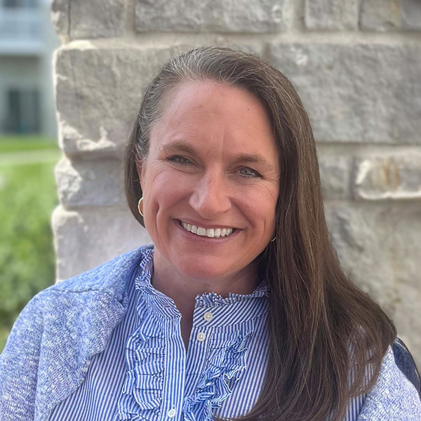 Kelly Flavin, Senior Living Coordinator at The Crestone Senior Living, smiling outdoors in front of a light stone wall. She is wearing a blue and white striped ruffled blouse with a blue knit cardigan.