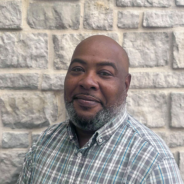 Pete Cooper, Culinary Director at The Crestone Senior Living, smiling in front of a light stone wall while wearing a gray and teal plaid button-down shirt.