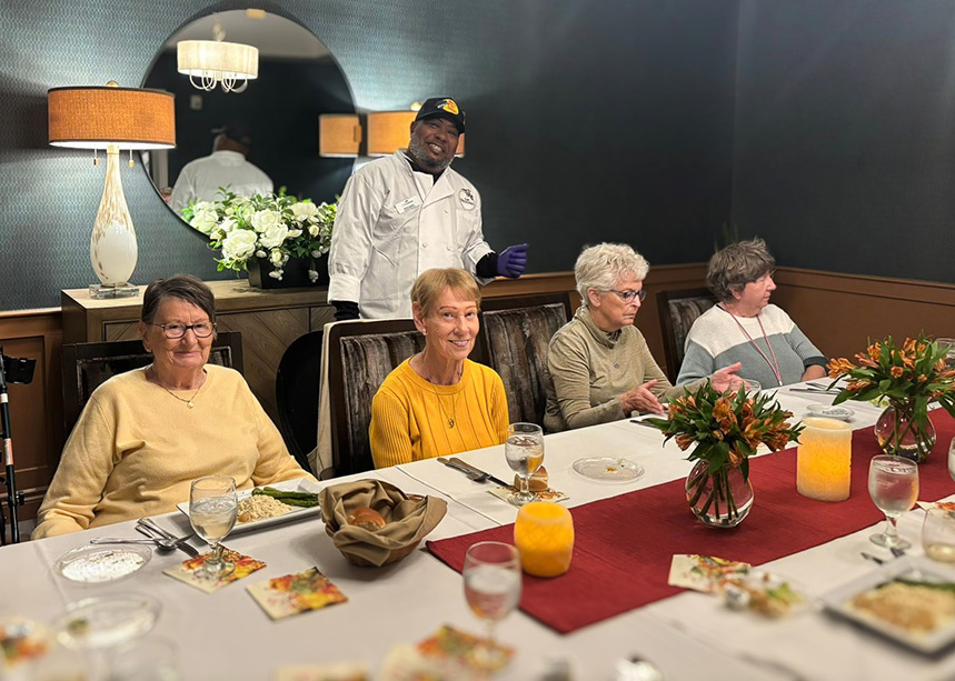 Senior women seated at a beautiful, elegantly decorated dining table, with chef smiling alongside them.
