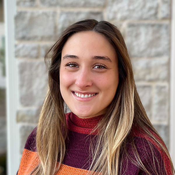 Kamryn Domenick, Memory Care Director at The Crestone Senior Living, smiling in a professional headshot, wearing a striped knit sweater, standing in front of a light stone exterior wall.