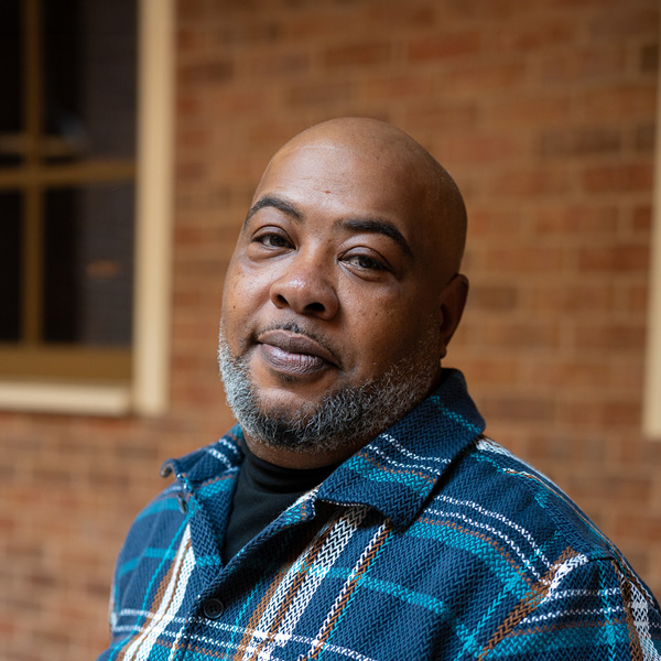 Pete Cooper, Culinary Director at The Crestone Senior Living, posing in a professional headshot, wearing a blue plaid shirt, with a softly blurred brick and window background.