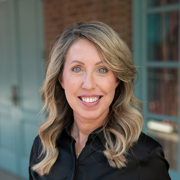 Jesse Alldredge, Executive Director at The Crestone Senior Living, smiling in a professional outdoor headshot with shoulder-length wavy blonde hair, bright blue eyes, and softly blurred brick and teal window details in the background.