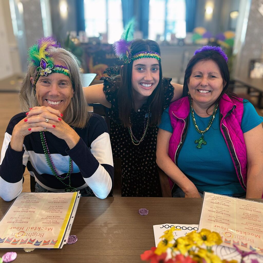 Three women smile widely, wearing brightly colored Mardi Gras themed accessories.