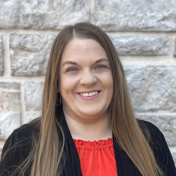 Brittney Cecil, Operations Coordinator at The Crestone Senior Living, smiling in a professional headshot with long straight light brown hair, hoop earrings, a red blouse, a black blazer, and a light gray stone wall background.