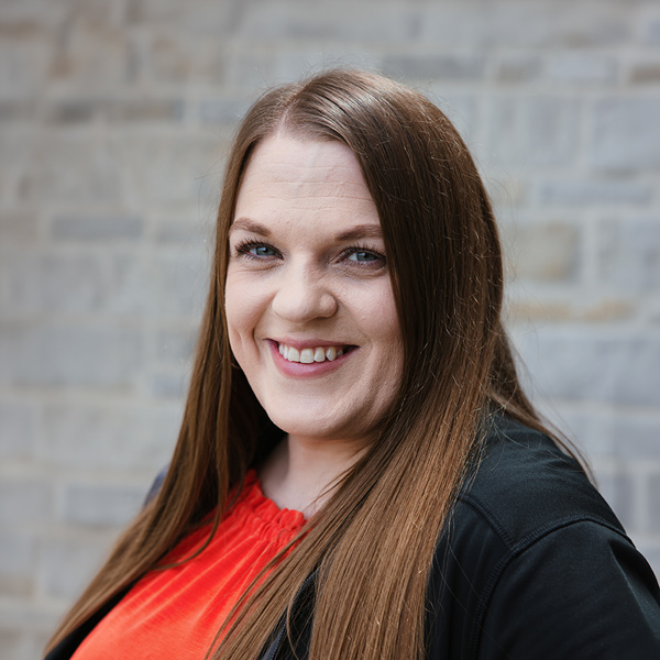 Brittney Cecil, Operations Coordinator at The Crestone Senior Living, smiling in a professional headshot turned slightly to the side, with long straight light brown hair, a bright red blouse, a black blazer, and a softly blurred light stone wall background.