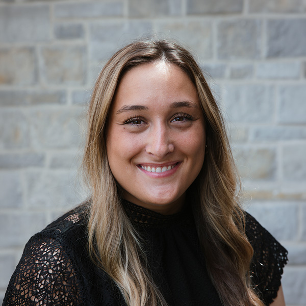 Kamryn Domenick, Memory Care Director at The Crestone Senior Living, smiling in a professional headshot with long softly waved highlighted hair, a black lace-sleeve blouse, and a softly blurred light stone wall background.