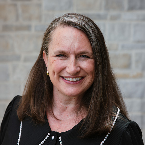 Kelly Flavin, Senior Living Coordinator at The Crestone Senior Living, smiling in a professional headshot with straight brown hair, a black blouse with pearl trim, small gold hoop earrings, and a softly blurred light stone wall background.