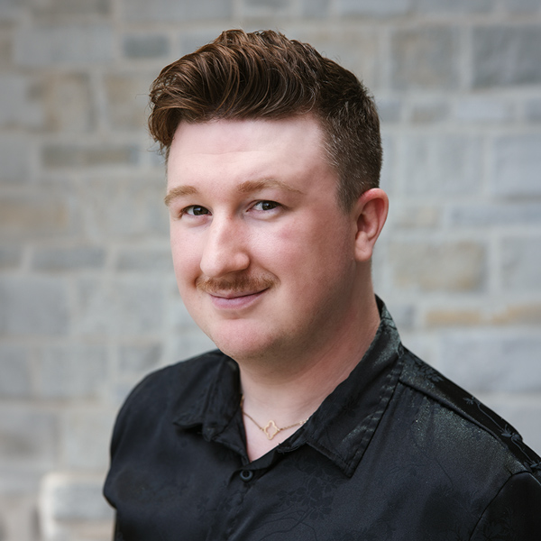 Payton Dishman, Resident Services Director at The Crestone Senior Living, looking at the camera in a professional headshot with short styled auburn hair, a mustache, a black patterned shirt, and a softly blurred light stone wall background.