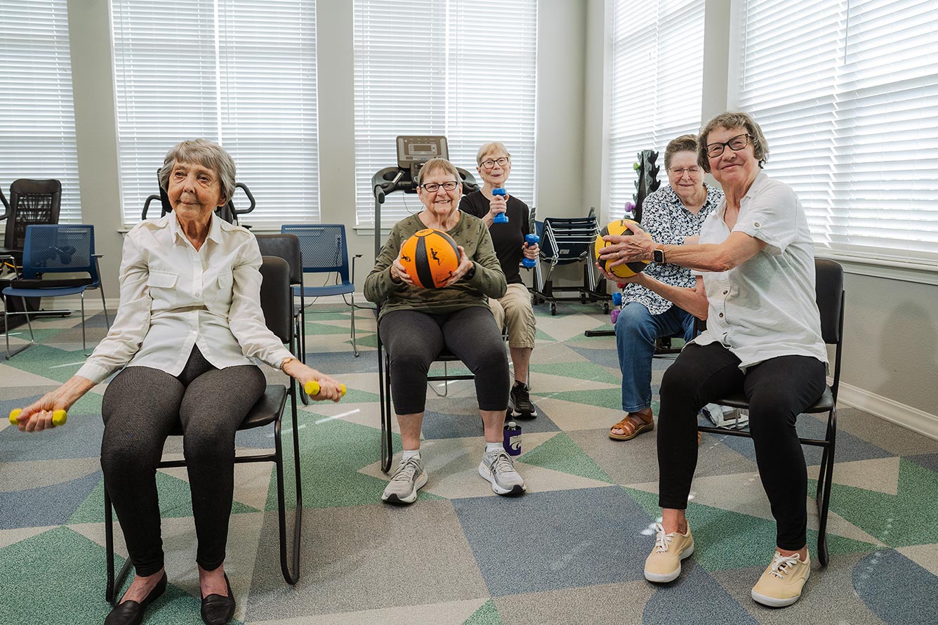 A group of senior residents hold small dumbbell weights or medicine balls while engaging in healthful chair exercises at The Crestone Senior Living to support their own wellness.