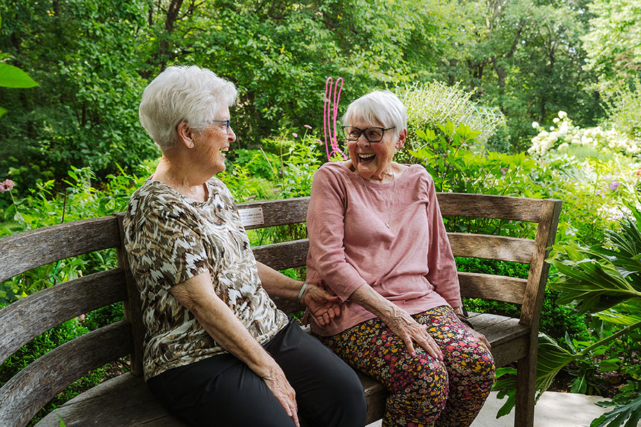 Two senior residents of The Crestone Senior living, chat and laugh together while enjoying a sunny day during a community outing to the local Arboretum.
