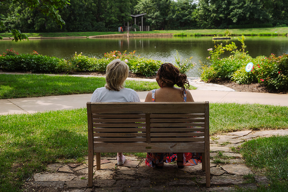 A senior resident and an employee sit on a bench together enjoying a pleasant view of the Arboretum's pond surrounded by greenery and flowers during a community outing with the residents of The Crestone Senior Living.