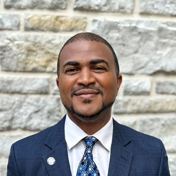 Demetrius Jones, Business Office Director at The Crestone Senior Living, looking at the camera in a professional headshot with short dark hair, a trimmed goatee, a navy suit, patterned tie, and a light stone wall background.